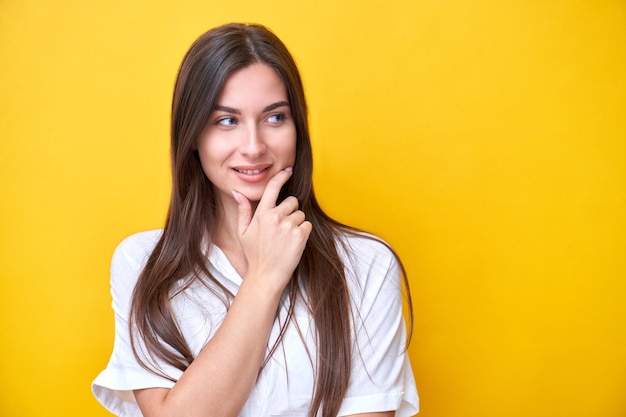 Ritratto di ragazza bruna sorridente tenendo il mento e pensando guarda di lato allo spazio di copia isolato su sfondo giallo