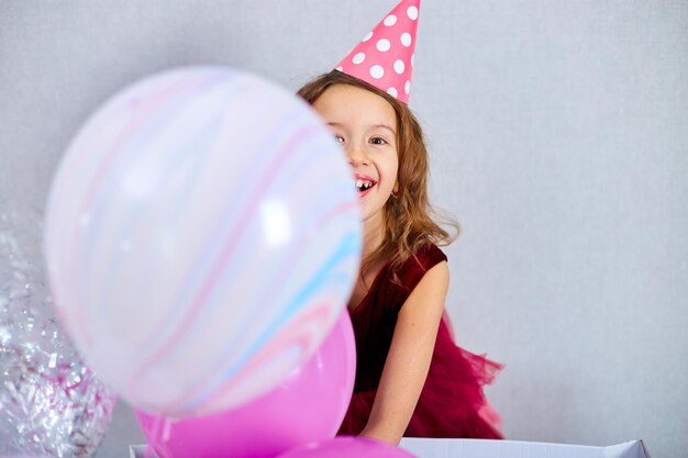 Ritratto di ragazza carina gioiosa in abito rosa e cappello gioca con palloncini a casa stelle filanti festa di compleanno Buon compleanno Celebrando