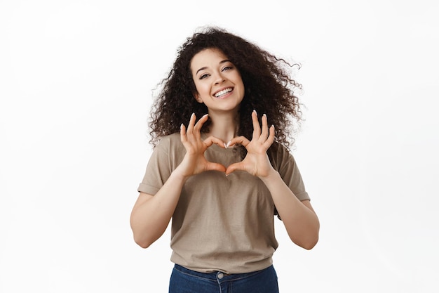 Ritratto di ragazza carina romantica smilig, mostrando il cuore ti amo segno e guardando la fotocamera, come te, in piedi su sfondo bianco