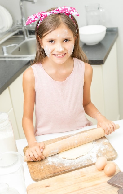 Ritratto di ragazza carina sorridente che rotola la pasta con il perno di legno sulla cucina