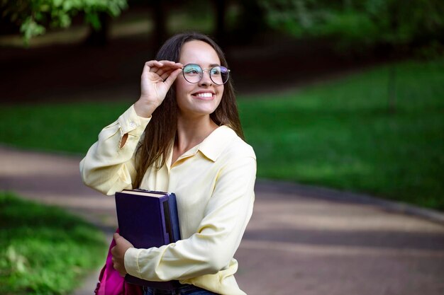 Ritratto di ragazza carina studentessa allegra che regola gli occhiali su un percorso del parco del campus