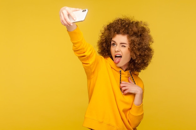 Ritratto di ragazza dai capelli ricci alla moda spensierata in felpa con cappuccio in stile urbano che si fa selfie e fa una smorfia con la lingua fuori divertendosi mentre fa foto vlog broadcast girato in studio isolato su sfondo giallo