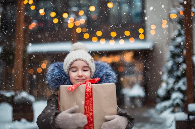 Ritratto di ragazza gioiosa con una confezione regalo per Natale in una strada cittadina in inverno con la neve in un mercato festivo con decorazioni e lucine Vestiti caldi cappello lavorato a maglia sciarpa e pelliccia Capodanno