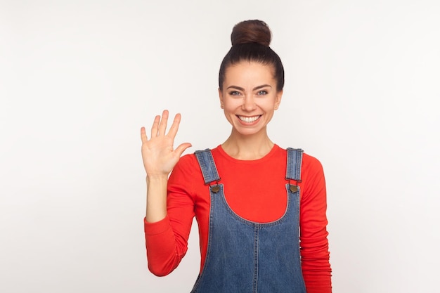 Ritratto di ragazza graziosa ed elegante con capelli chignon in tuta di jeans che fa un gesto di saluto dolce, accogliendo con un sorriso a trentadue denti, agitando la mano e salutando. tiro in studio indoor isolato su sfondo bianco