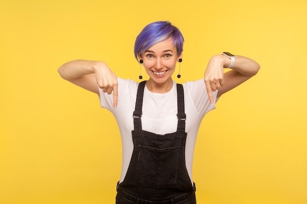 Ritratto di ragazza hipster sorridente felice alla moda con i capelli corti viola in tuta rivolta verso il basso che mostra lo spazio per la pubblicità, il tuo testo promozionale. isolato su sfondo giallo girato in studio