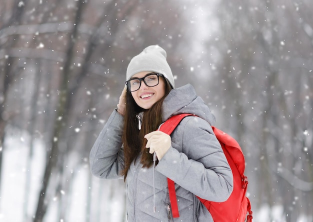 Ritratto di ragazza hipster turistica con gli occhiali che indossa una giacca grigia che cammina con uno zaino rosa nella foresta fredda invernale