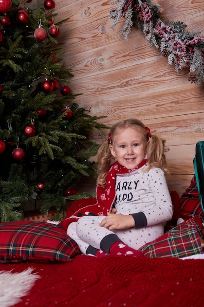 Ritratto di ragazza in pigiama seduta sul letto sotto l'albero di Natale con regali in scatole regalo.