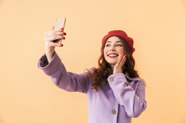 Ritratto di ragazza positiva 20s in cappotto e cappello che cattura foto selfie sul telefono cellulare, mentre in piedi isolato