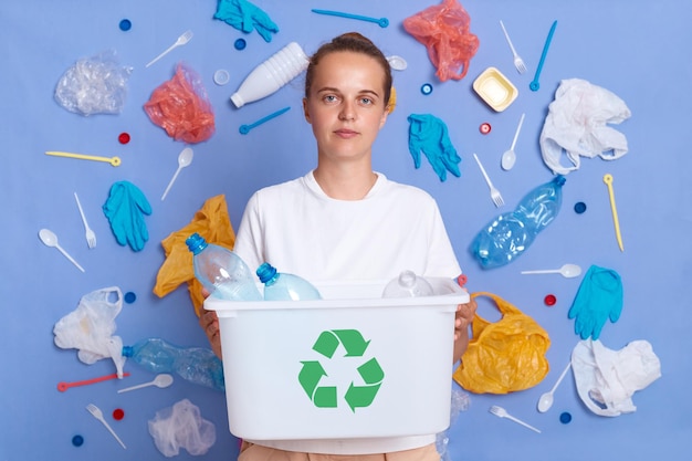 Ritratto di ragazza seria concentrata con capelli scuri che tiene scatola di bottiglie di plastica e riciclaggio simbolo verde smistamento spazzatura risparmio ecologia in posa su sfondo blu con spazzatura