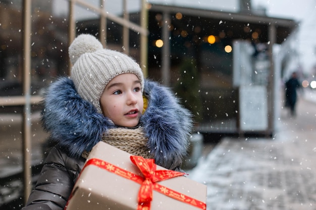 Ritratto di ragazza sorpresa con una confezione regalo per Natale in una strada cittadina in inverno con la neve in un mercato festivo con decorazioni e luci. Vestiti caldi, berretto lavorato a maglia, sciarpa e pelliccia. Copia spazio