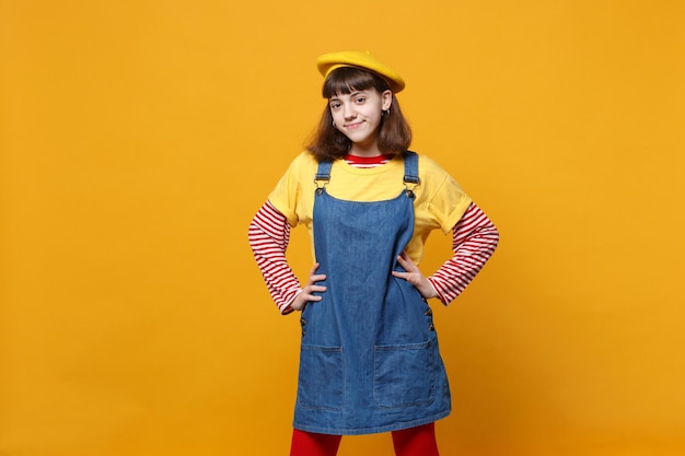 Ritratto di ragazza sorridente adolescente in berretto francese, prendisole in denim in piedi con le braccia sui fianchi in vita isolato su sfondo giallo muro. Persone sincere emozioni, concetto di stile di vita. Mock up spazio di copia.