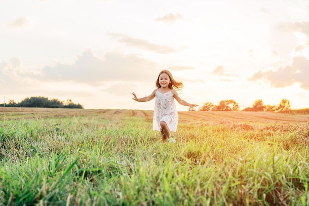 Ritratto di ragazza sorridente che gioca a saltare e correre sull'erba campi di fieno percorsi di erba secca al tramonto Mani d'ondeggiamento Foresta su sfondo luminoso Cielo soleggiato nuvoloso Tempo di fienagione