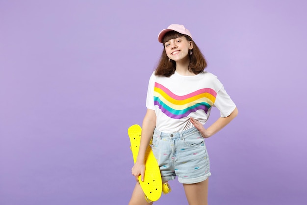 Ritratto di ragazza teenager sorridente allegra in abiti vivaci che tiene skateboard giallo isolato su sfondo muro pastello viola in studio. Persone sincere emozioni, concetto di stile di vita. Mock up spazio di copia.