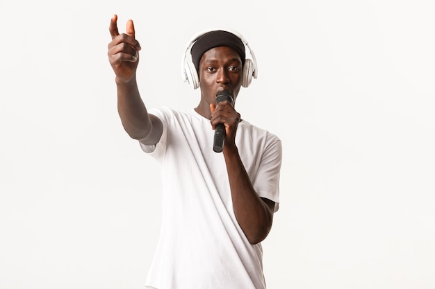 Ritratto di ragazzo afro-americano impertinente e freddo in cuffie wireless, cantando nel microfono