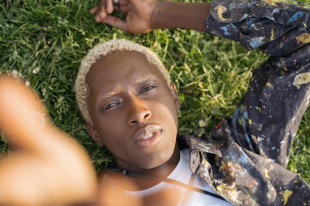 Ritratto di ragazzo afroamericano con capelli alla moda prendendo selfie guardando la fotocamera sdraiato sull'erba