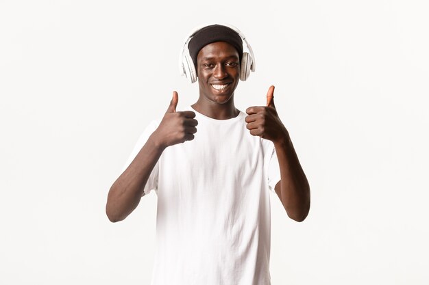 Ritratto di ragazzo afroamericano soddisfatto e sorridente che gode della musica impressionante, mostrando il pollice in su in approvazione, ascoltando canzoni in cuffia.