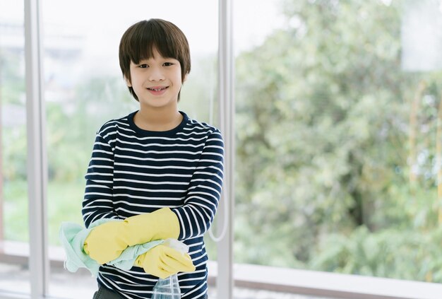 Ritratto di ragazzo asiatico sorridente con flacone spray e straccio che indossa guanti di gomma e guarda la fotocamera a casa Ragazzo che aiuta a pulire la casa Pulizia dei bambini e concetto di aiuto per le pulizie