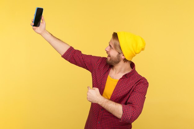 Ritratto di ragazzo barbuto hipster con cappello a cuffia e camicia a scacchi che parla in videochiamata, conversazione online sul telefono cellulare, prendendo selfie. Studio indoor girato isolato su sfondo giallo.