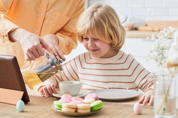 Ritratto di ragazzo sorridente sveglio che gode della colazione in cucina e guarda i cartoni animati tramite il computer portatile