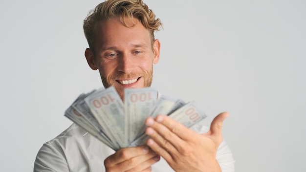 Ritratto di ricco uomo d'affari barbuto biondo che sembra felice mostrando mazzetta di soldi sulla fotocamera e sorridente su sfondo bianco