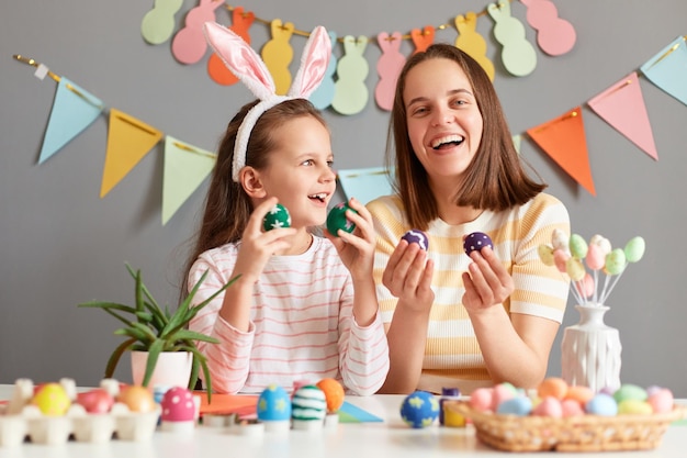 Ritratto di ridere madre e figlia estremamente felici che indossano orecchie da coniglio che tengono uova colorate che celebrano la Pasqua a casa in posa contro il muro decorato grigio