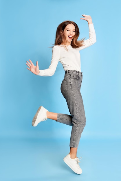 Ritratto di salto positivo di giovane bella ragazza in camicetta bianca e jeans in posa su studio blu