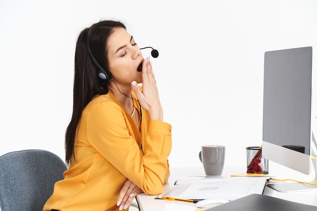 Ritratto di sbadiglio assistente hotline donna che indossa il microfono auricolare parlando con il cliente al telefono mentre si lavora in ufficio
