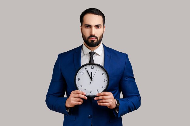 Ritratto di serio uomo barbuto prepotente paura di essere in ritardo tenendo in mano orologio da parete scadenza puntualità indossando abito stile ufficiale Indoor studio shot isolato su sfondo grigio