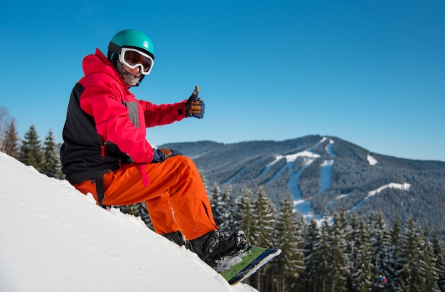 Ritratto di snowboarder maschio seduto, rilassante sul pendio nevoso in inverno stazione sciistica in montagna, mostrando i pollici fino alla telecamera