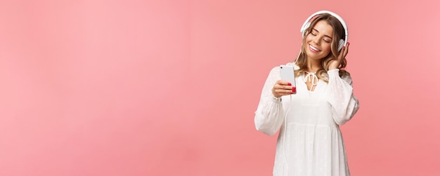 Ritratto di sognante ragazza bionda carina e tenera in abito bianco che tiene smartphone ascolta musica in cuffia sorridendo al display del telefono cellulare come playlist di selezione per sfondo rosa umore primaverile