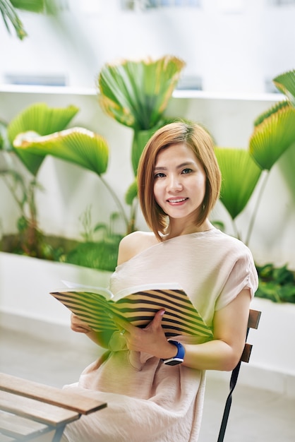 Ritratto di sorridente attraente giovane donna vietnamita seduto al tavolo del bar con il libro aperto nelle mani