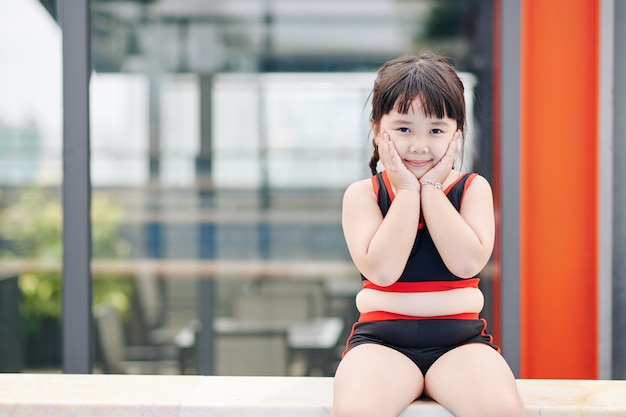 Ritratto di sorridente bambina asiatica seduta sul bordo della piscina, toccando le guance e guardando