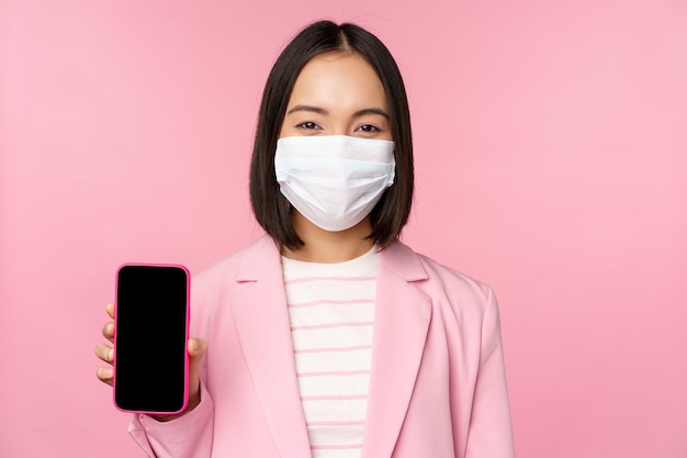 Ritratto di sorridente commessa coreana in tuta medica con maschera facciale che mostra lo schermo dello smartphone in piedi su sfondo rosa