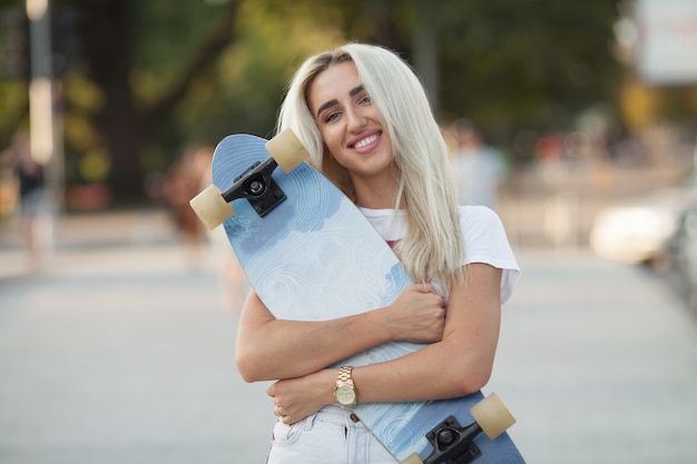 Ritratto di sorridente giovane guidatore di skateboard femminile che tiene il suo skateboard. Donna con tavola da pattinaggio a skate park guardando la fotocamera all'esterno.