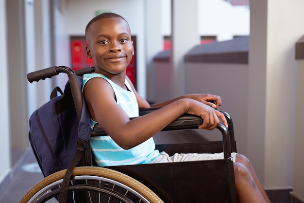 Ritratto di sorridente scolaro afroamericano disabile seduto in sedia a rotelle nel corridoio della scuola