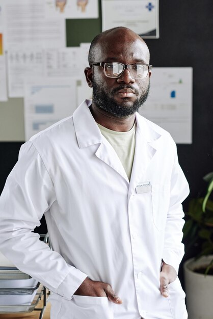 Ritratto di specialista medico africano in occhiali e camice da laboratorio in piedi in ufficio e guardando