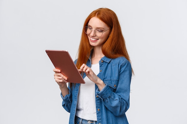 Ritratto di spensierata donna europea spensierata con i capelli rossi con gli occhiali, guardando divertito e divertito con tavoletta digitale, sorridendo felice, soddisfatto del risultato, in piedi