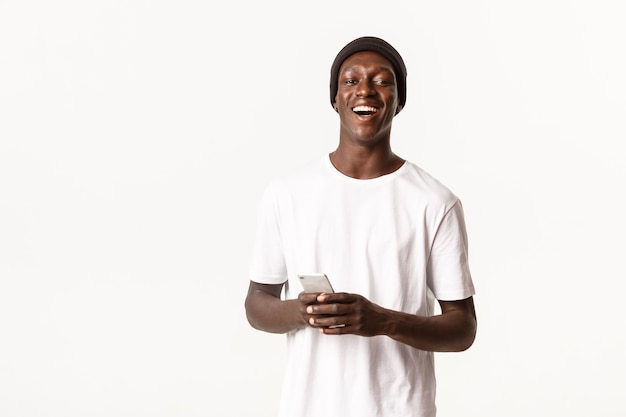 Ritratto di spensierato attraente ragazzo afro-americano in beanie ridendo e sorridendo, utilizzando l'applicazione del telefono cellulare