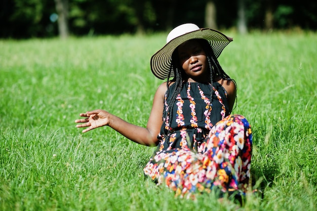 Ritratto di splendida donna afroamericana anni '20 in cappello estivo seduto all'erba verde nel parco.