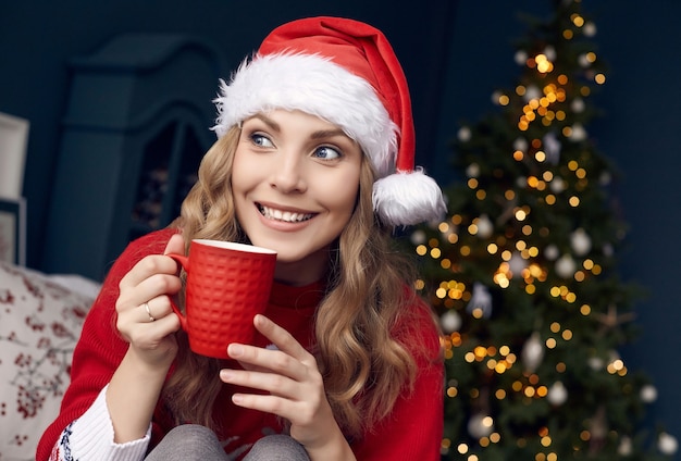 Ritratto di splendida donna bionda in maglione rosso e cappello della Santa con una tazza di caffè in posa all'interno decorato di Natale.