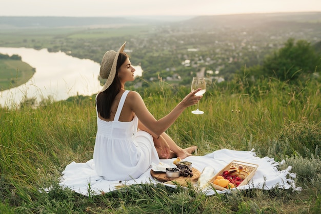 Ritratto di splendida donna bruna in abito bianco e cappello di paglia con vino sulla collina con un bellissimo paesaggio