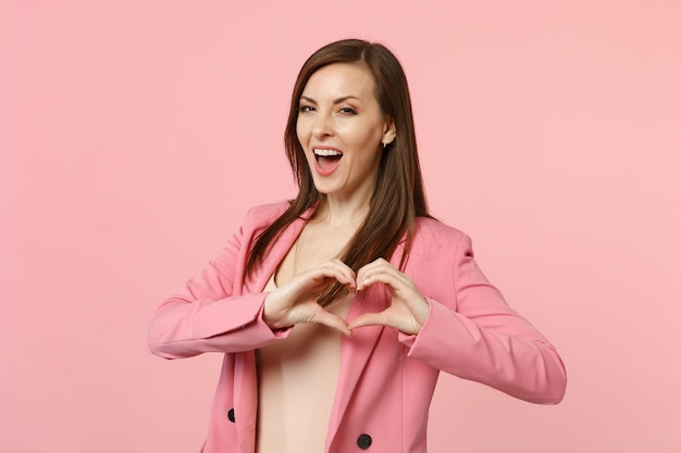 Ritratto di splendida giovane donna allegra in giacca che mostra forma cuore con le mani isolate su sfondo rosa pastello parete in studio. Persone sincere emozioni, concetto di stile di vita. Mock up spazio di copia.