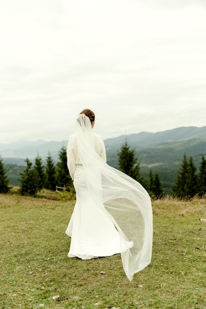 Ritratto di sposa Una giovane ragazza in un abito da sposa bianco e con un mazzo di fiori e verde in mano sullo sfondo di montagne e foreste al tramonto