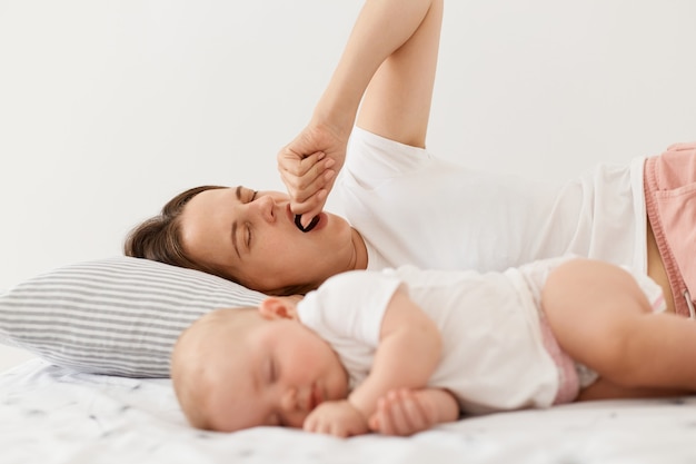 Ritratto di stanca madre esausta sdraiata sul letto e sbadigliando, coprendo la bocca aperta con il pugno, si sveglia presto mentre la sua bambina dorme, posando a casa in una stanza luminosa.