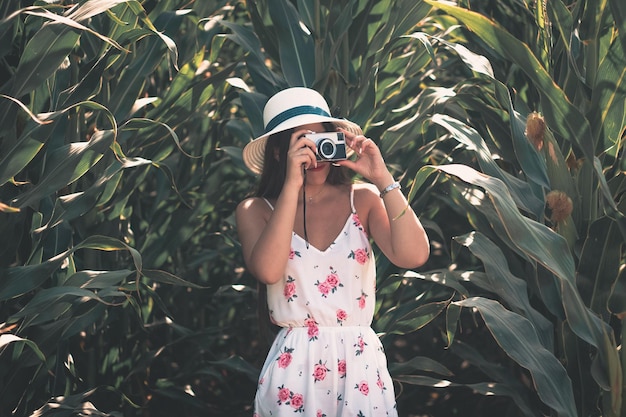 Ritratto di stile di vita all'aperto di una giovane ragazza bruna che scatta foto con una macchina fotografica d'epoca che indossa un abito estivo a fiori bianchi e un cappello da sole