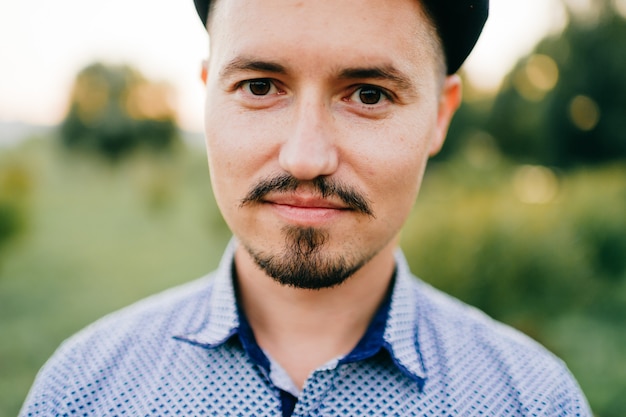 Ritratto di stile di vita del primo piano dell'uomo kazako bello che posa di estate all'aperto sul fondo astratto della natura. Maschio felice sorridente con i baffi e la protezione da portare della barba. Ragazzo adulto carismatico in maglietta blu