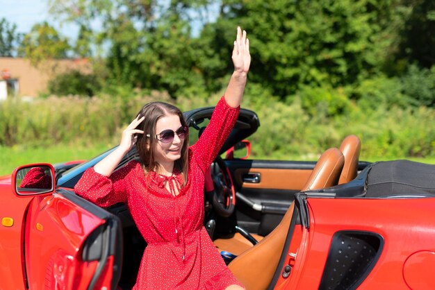 Ritratto di stile di vita di bella donna spensierata in vestito rosso e occhiali da sole che si siedono sul cabriolet rosso