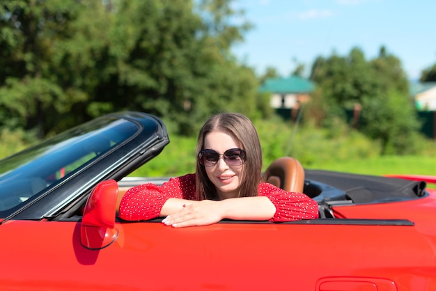 Ritratto di stile di vita di bella donna spensierata in vestito rosso e occhiali da sole che si siedono sul cabriolet rosso