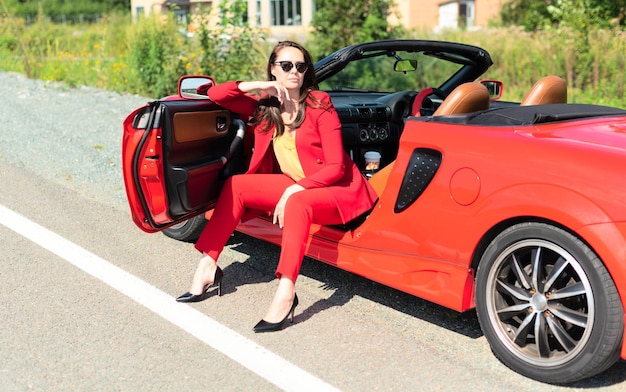 Ritratto di stile di vita di una bella donna spensierata in occhiali da sole e vestito rosso che si siede sul cabrio rosso