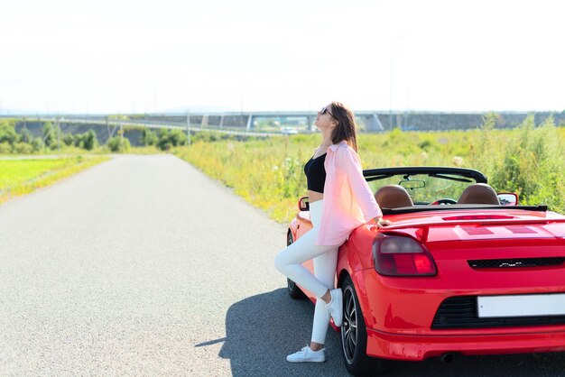 Ritratto di stile di vita di una bella donna spensierata vestita casualmente in piedi vicino a un'auto cabriolet rossa e sorridente Viaggio su strada godendo del concetto di libertà